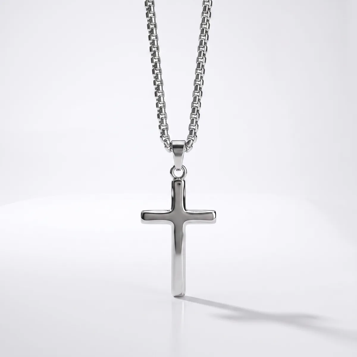 A polished silver cross pendant hangs from a matching chain against a white background, casting a soft shadow.