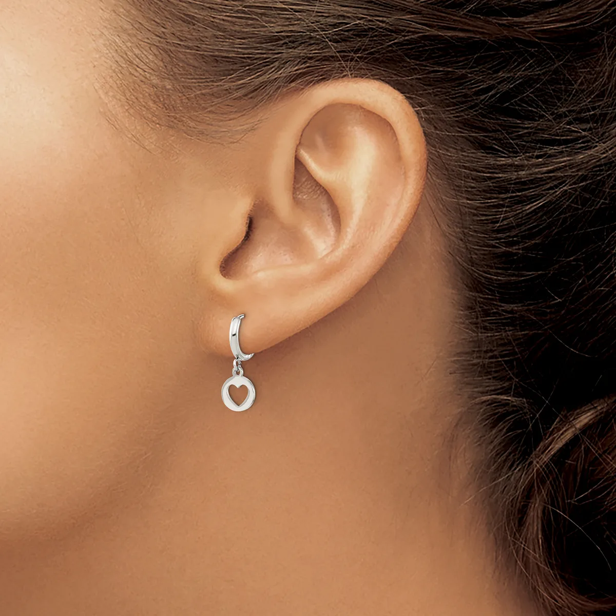 A woman wearing a silver heart-shaped dangle earring with a hoop post, shown from the side view of her ear.