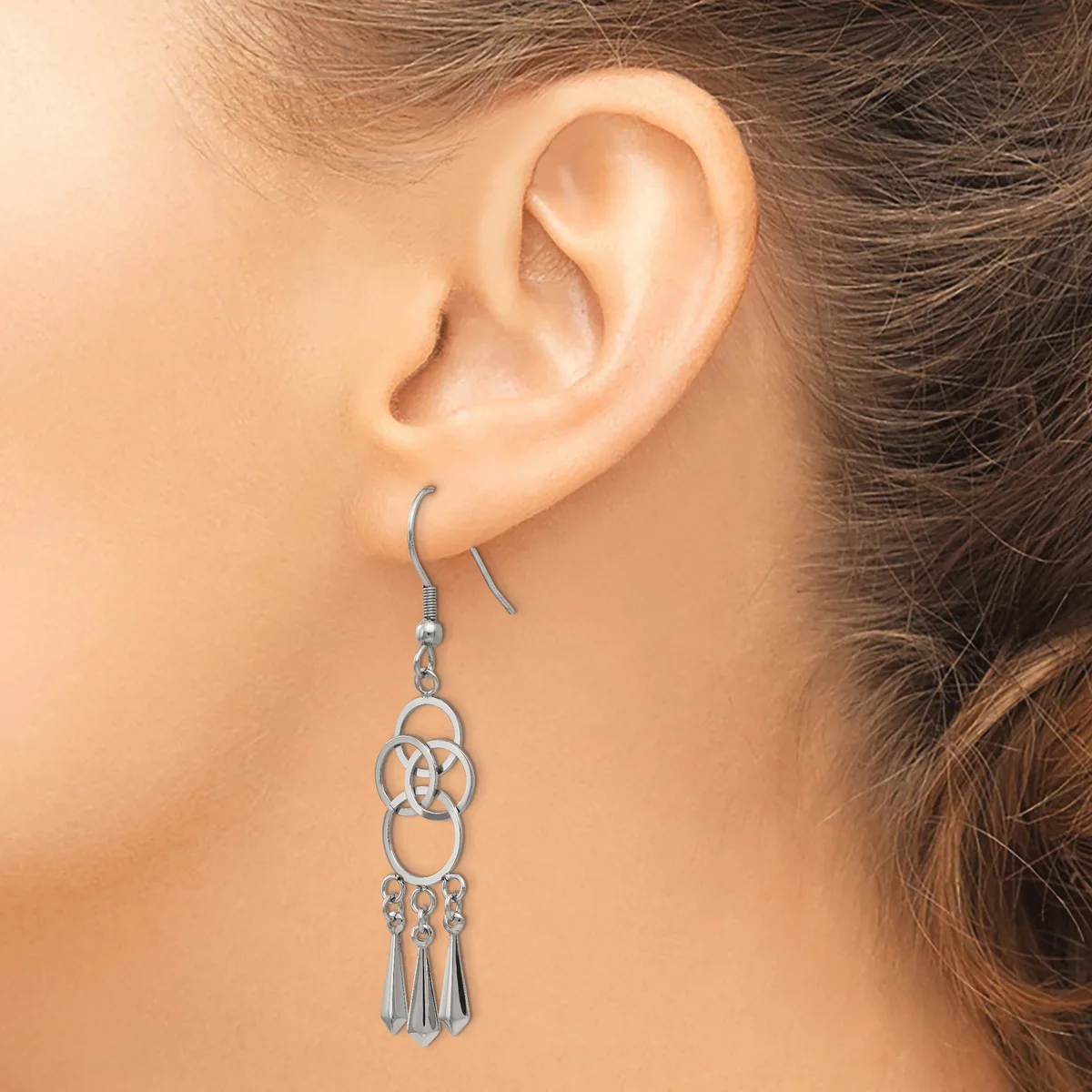 A silver-toned dangle earring with interlocking circular designs and tassel details hangs from a fish hook, worn on a person's ear.