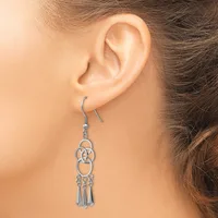A silver-toned dangle earring with interlocking circular designs and tassel details hangs from a fish hook, worn on a person's ear.