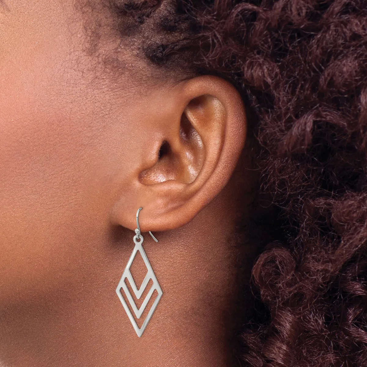 A silver geometric diamond-shaped dangle earring with nested chevron cutouts hangs from an ear lobe.