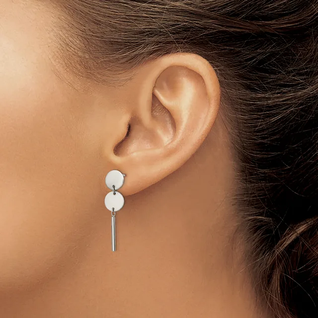 A woman wearing a pair of silver drop earrings with two white spheres stacked vertically and a thin bar hanging below, shown from the side view of her ear and neck.