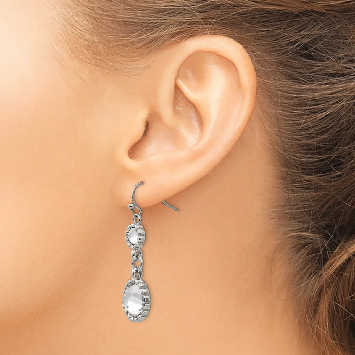 A woman wears a pair of dangling earrings with oval-shaped clear gemstones set in silver-toned metal, suspended from hook-style ear wires.