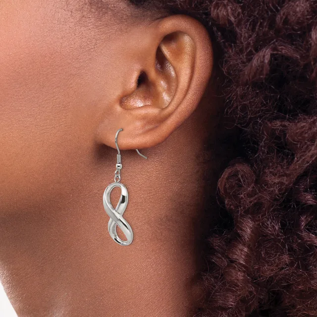 A silver infinity symbol earring hangs from a fish hook wire on a person's earlobe, shown in profile view against their neck and curled dark hair.