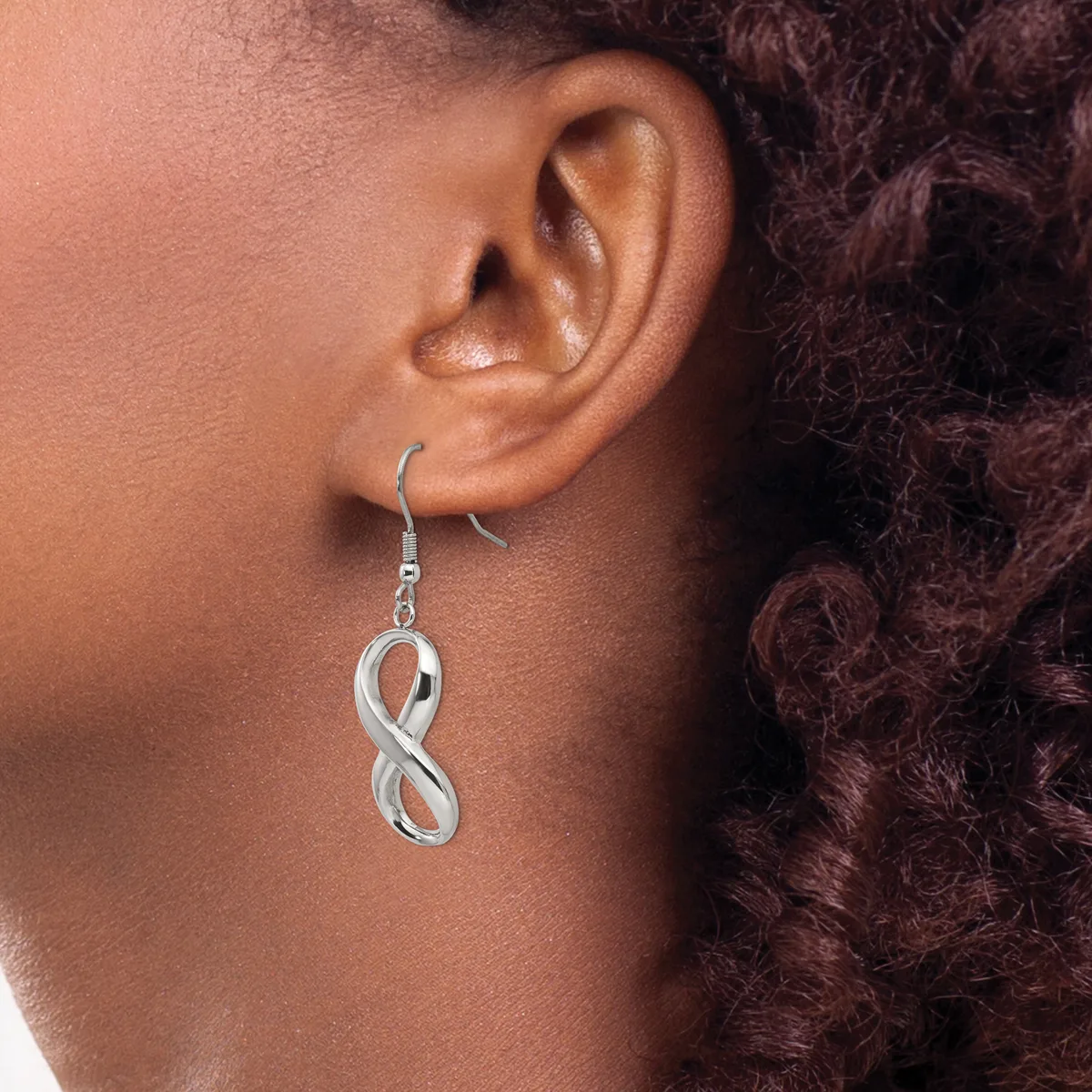 A silver infinity symbol earring hangs from a fish hook wire on a person's earlobe, shown in profile view against their neck and curled dark hair.