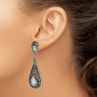 A woman wearing ornate silver teardrop earrings with white stone centers and detailed black patterning, shown from the side profile against her ear.