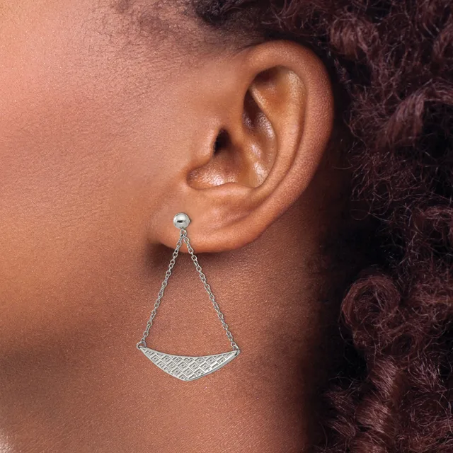A person wearing a silver triangular pendant earring with textured detailing on the lower section, suspended from a delicate chain and stud post.
