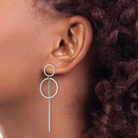Silver-toned drop earrings with circular geometric designs hanging from a person's ears, shown in profile against their shoulder and dark curly hair.