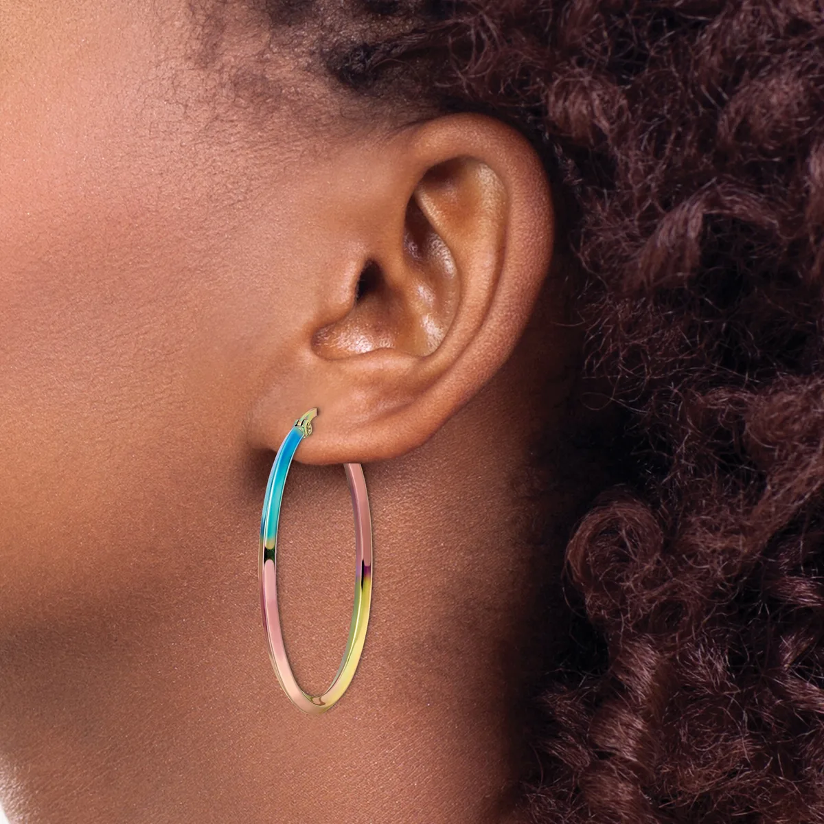 A person wearing a pair of large oval hoop earrings with a gradient color finish transitioning from blue to pink to yellow.