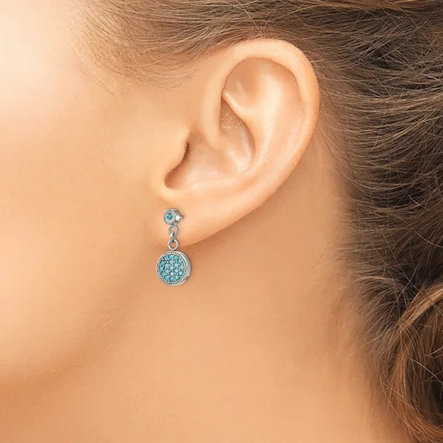 A woman wearing a dangling earring with a small round stud at the top connected to a larger circular pendant studded with light blue stones.