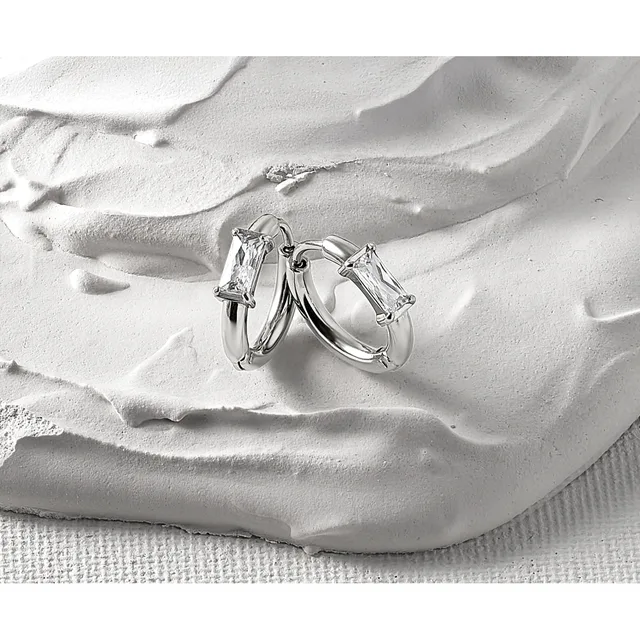 Two silver hoop earrings with rectangular clear crystal stones displayed on a textured white sculptural hand form against a light fabric background.