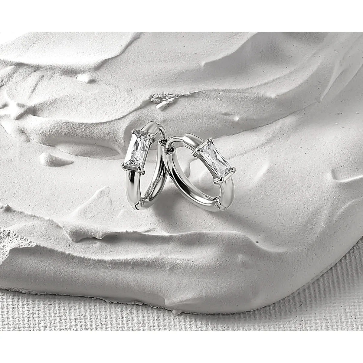 Two silver hoop earrings with rectangular clear crystal stones displayed on a textured white sculptural hand form against a light fabric background.