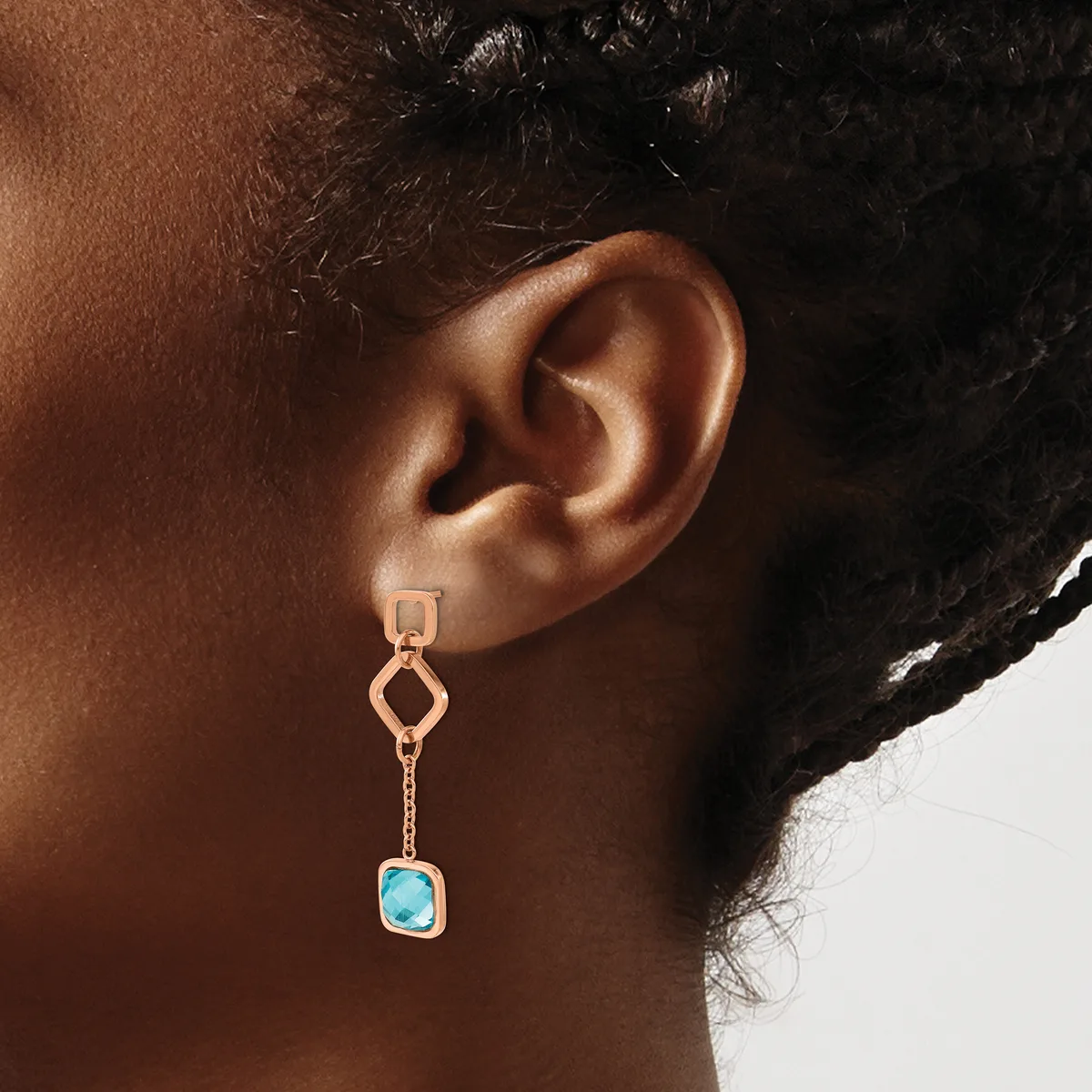 A person wearing a rose gold drop earring with a geometric upper link, delicate chain, and a cushion-cut turquoise stone in a bezel setting.