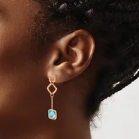 A person wearing a rose gold drop earring with a geometric upper link, delicate chain, and a cushion-cut turquoise stone in a bezel setting.