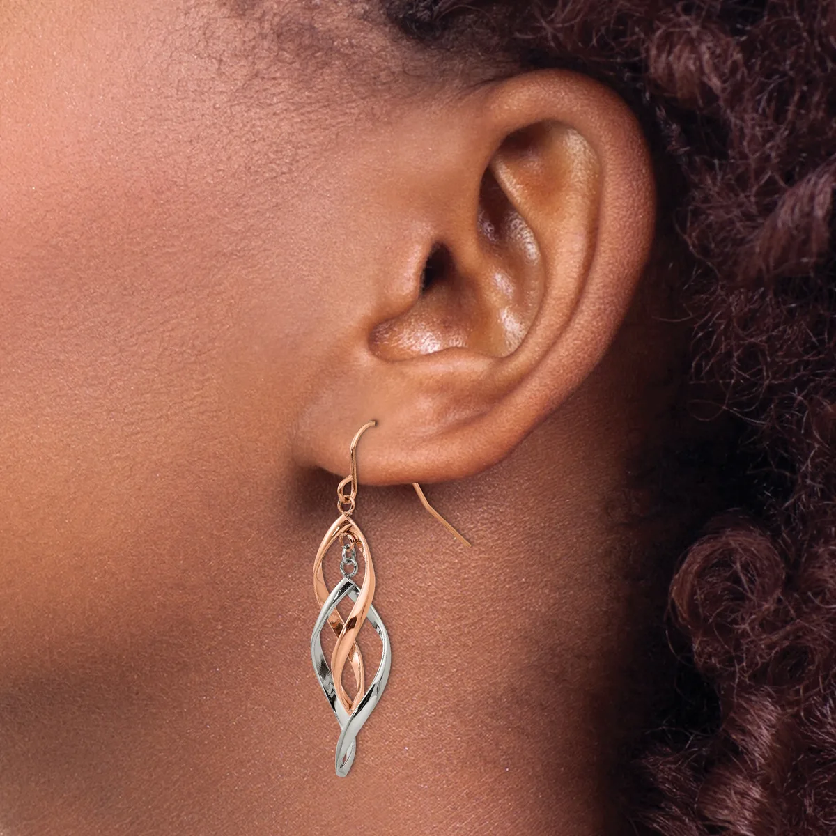 A person wearing a twisted drop earring in silver and rose gold tones hanging from a fish hook ear wire.