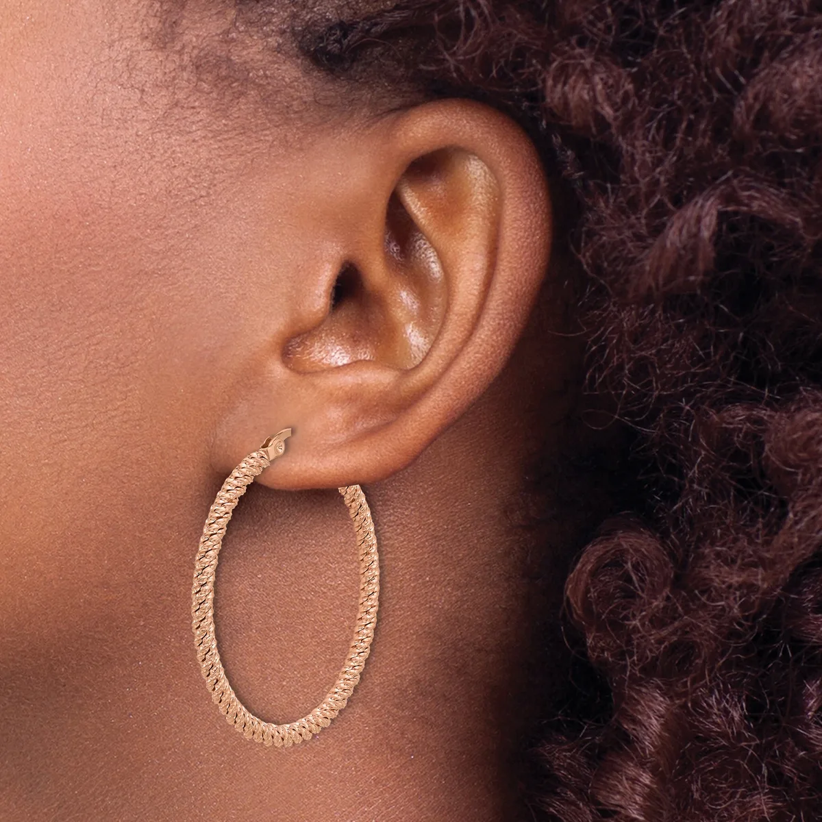 Close-up side view of a person's ear wearing a textured oval hoop earring in gold-toned metal.