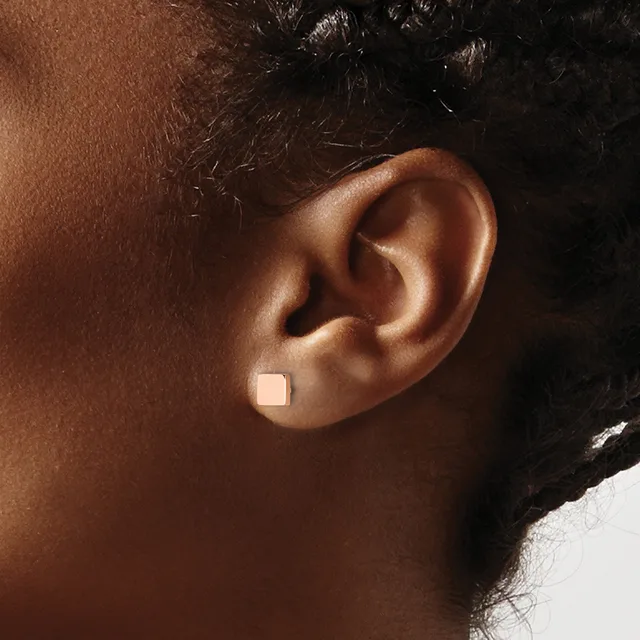 A person with dark skin and black hair wears a small square silver stud earring on their earlobe.