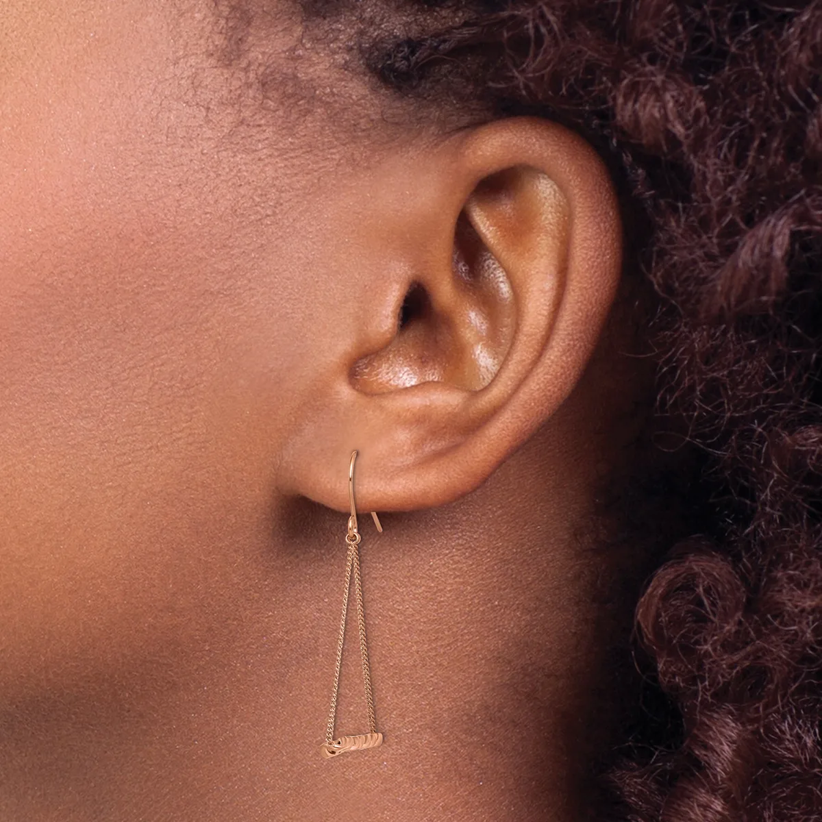 A person wearing a long silver-toned dangle earring with a rectangular pendant, shown in profile view against their ear and dark curled hair.