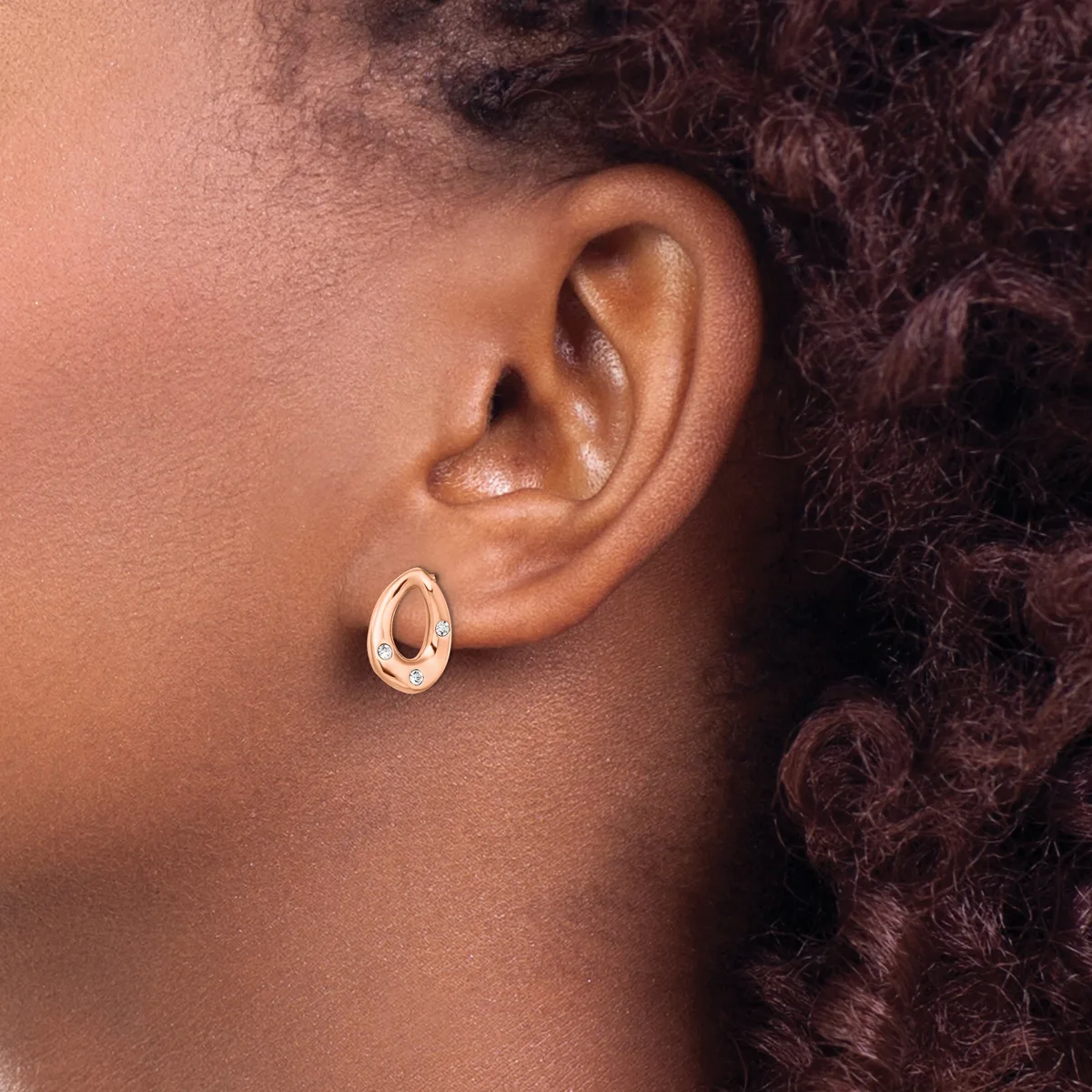 A person wearing a polished rose gold oval hoop earring, shown in profile view against their ear and dark curled hair.