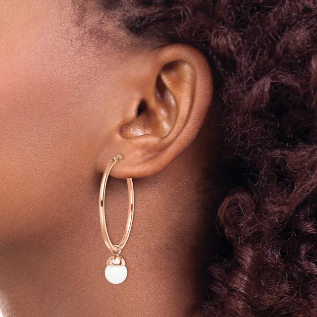 A person wearing a rose gold hoop earring with a small white circular pendant hanging from it.