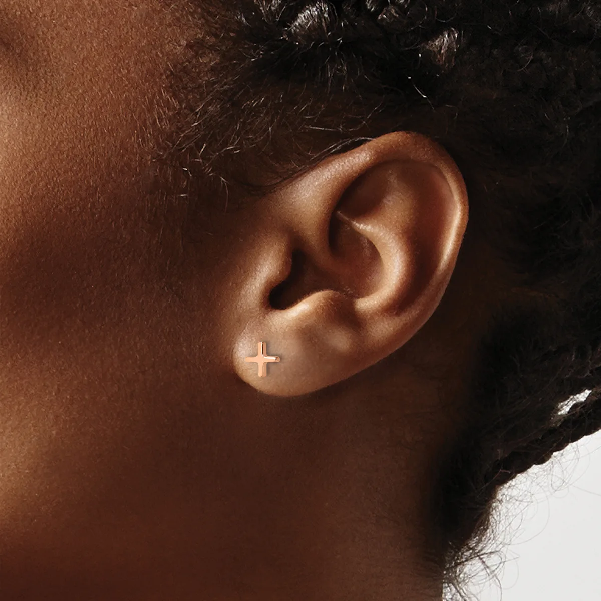 A small cross-shaped stud earring in white metal worn on the earlobe of a person with brown skin and dark hair.