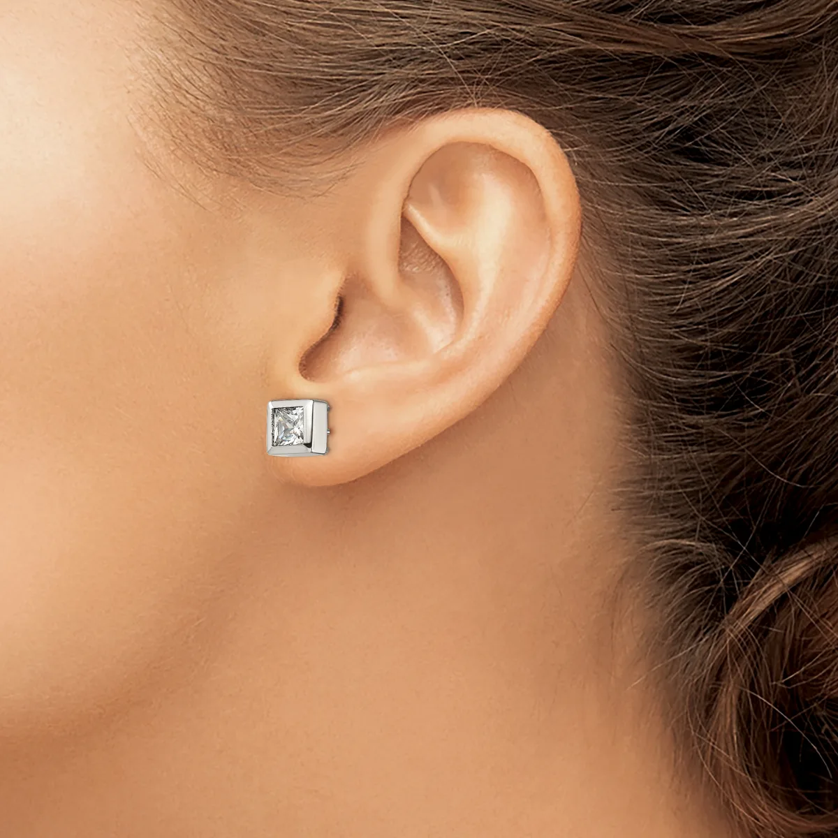 A person wearing a square stud earring with a clear stone set in a silver-toned metal setting, shown on their earlobe.
