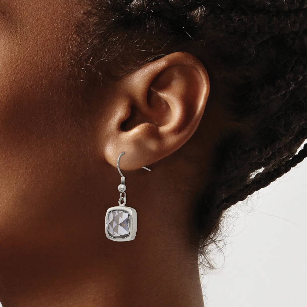 A close-up of a person's ear wearing a square silver dangle earring with a geometric pattern, suspended from a curved hook.