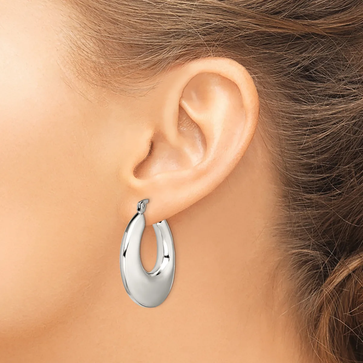 A woman wearing a polished silver-toned oval hoop earring in her pierced earlobe, shown from the side view of her head and neck.