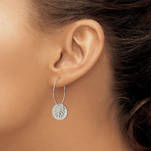 A person wearing silver hoop earrings with circular pendant charms stamped with leaf designs, shown from the side view of their ear.