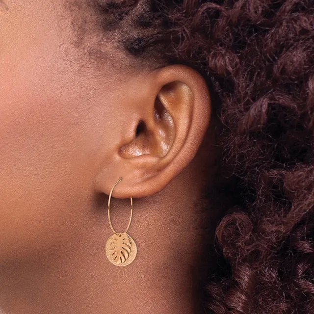 A person with dark curly hair wears a gold hoop earring with a dangling circular pendant stamped with a leaf design.