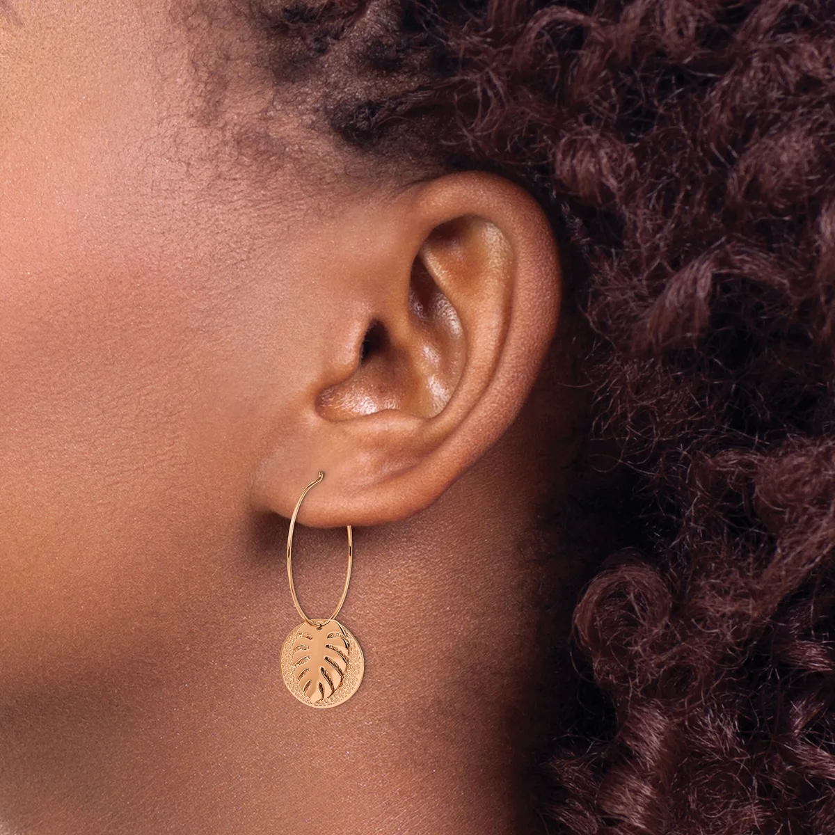 A person with dark curly hair wears a gold hoop earring with a dangling circular pendant stamped with a leaf design.