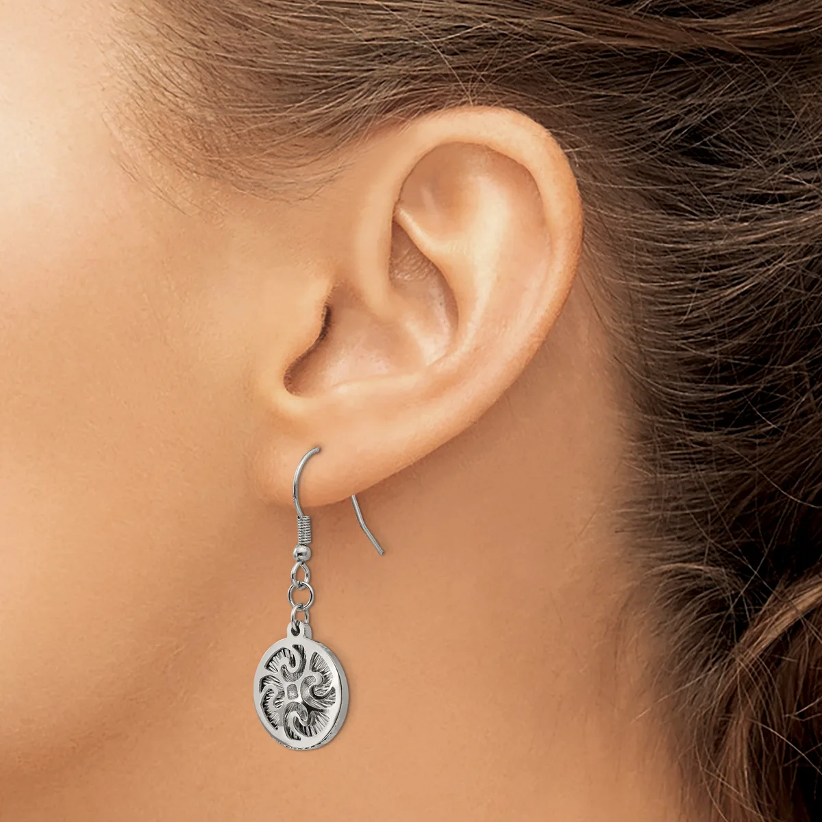 A person wearing a silver dangle earring with an oval pendant featuring an ornate floral design, shown from the side view of their ear.