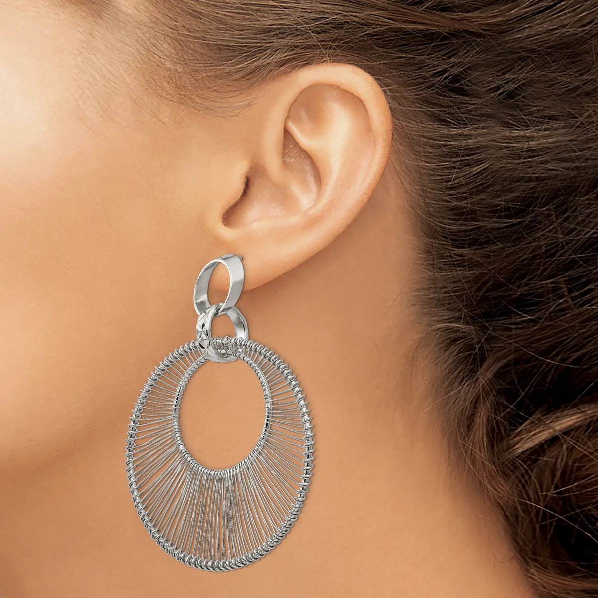 A woman wearing a large silver oval dangle earring with radiating wire details, shown from the side view against her ear.