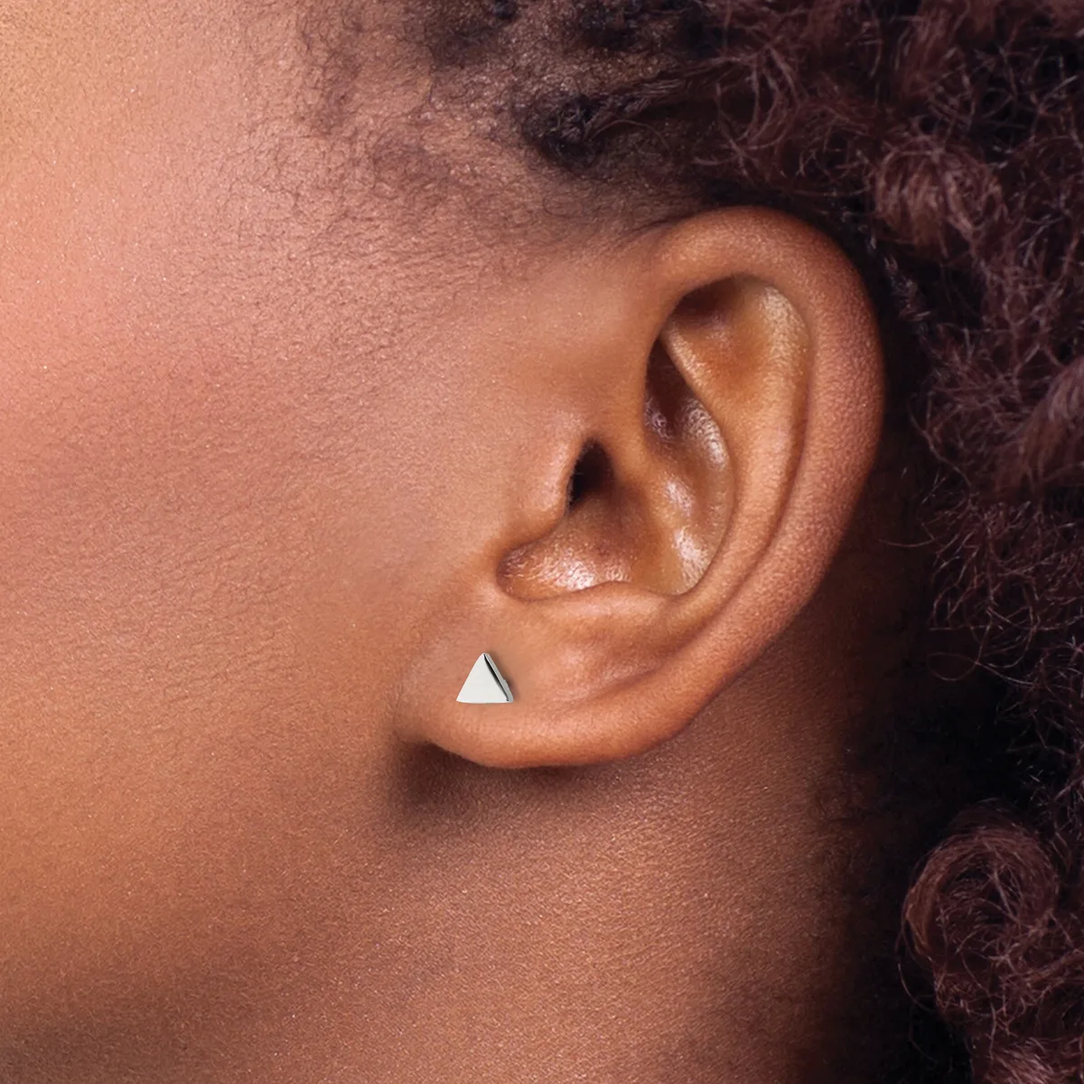 A small triangular silver stud earring worn in the lower lobe of a person with deep skin tone and textured dark hair.