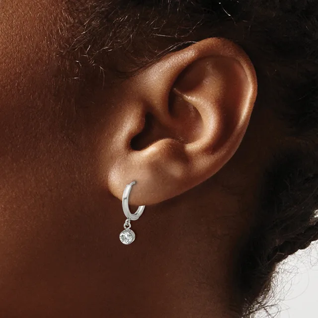 A silver hoop earring with a small round diamond pendant hangs from the earlobe of a person with brown skin and dark hair.