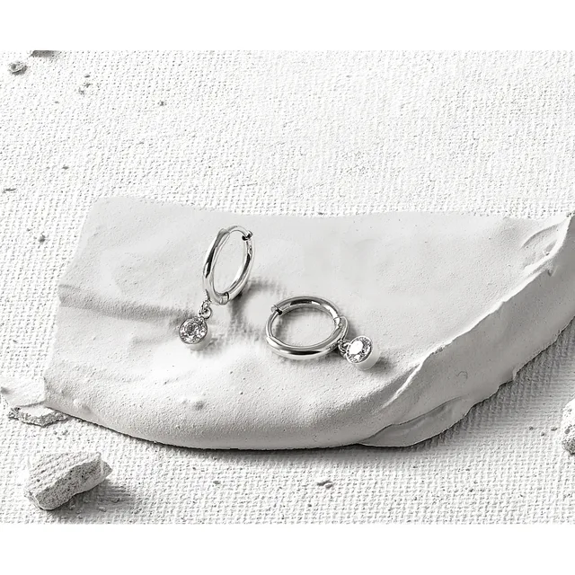Two silver hoop earrings with dangling round cubic zirconia stones displayed on a white textured fabric with a small stone nearby.
