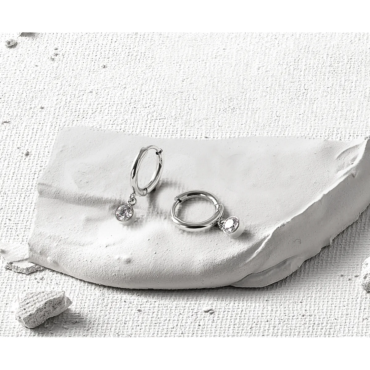 Two silver hoop earrings with dangling round cubic zirconia stones displayed on a white textured fabric with a small stone nearby.