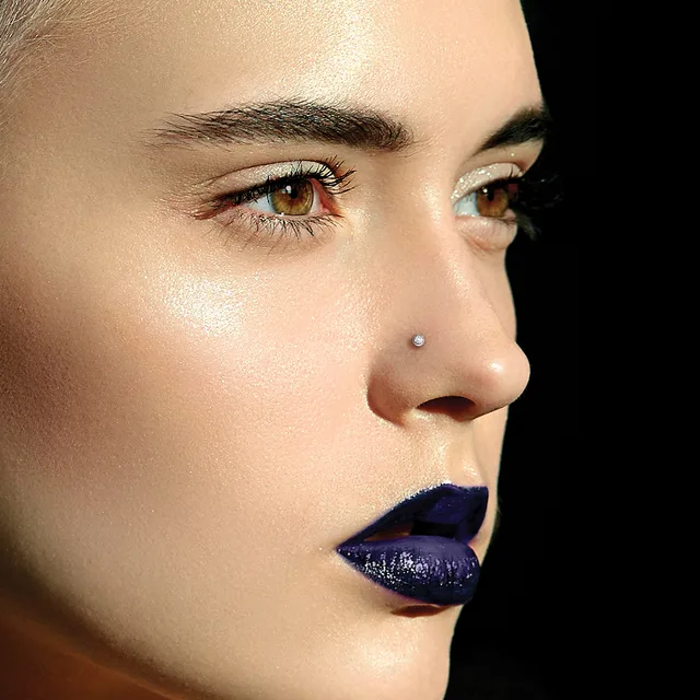 A person's face in profile against a black background, wearing dark blue lipstick, with a nose stud, defined eyebrows, and glowing skin.