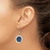 A woman wearing a dangling earring with a circular silver frame enclosing a dark blue glittery stone, suspended from a silver hook.