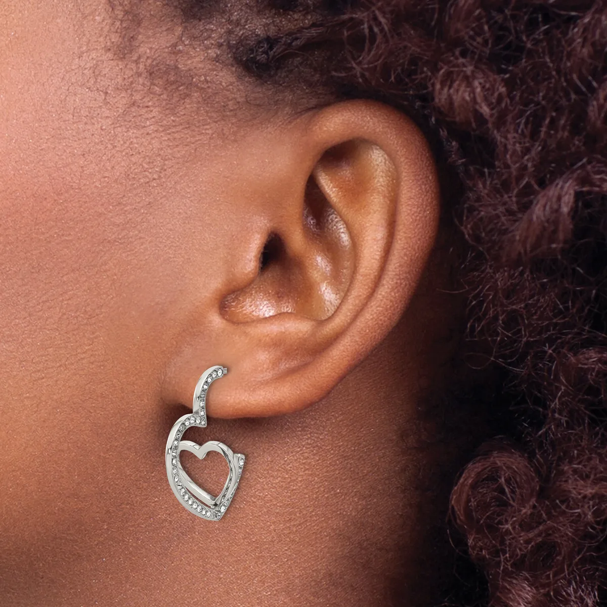 A silver heart-shaped earring with diamond or crystal embellishments hangs from a pierced earlobe against skin with dark curled hair visible in the background.