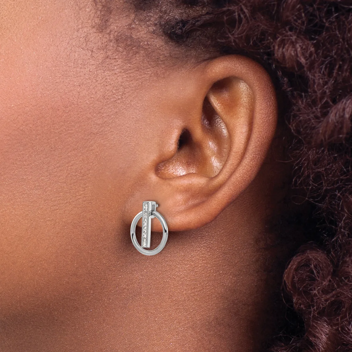 A silver circular drop earring with a vertical bar detail worn on the earlobe of a person with medium skin tone and dark curly hair.