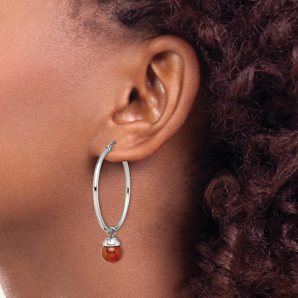 A person wearing a silver hoop earring with a dangling polished red stone pendant, shown from the side view against their ear.