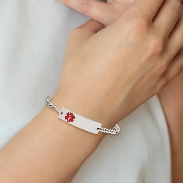 A person wearing a white medical alert bracelet with a red medical symbol on the ID plate, connected to silver-toned chain links on each side.
