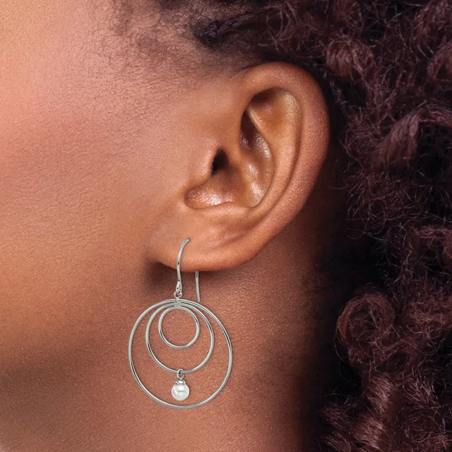 A person wearing a silver wire dangle earring with three concentric circular loops and a small pearl hanging from the innermost loop.