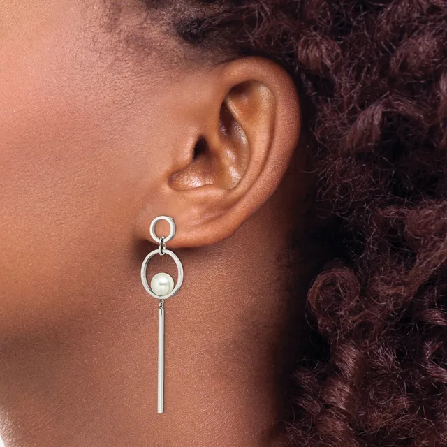 A person wearing a long silver dangle earring with circular loops at the top and a pearl accent, shown from the side view against their ear.
