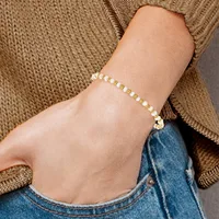A person wearing a beige knit sweater and blue jeans displays a beaded bracelet on their wrist made of alternating white and yellow round beads with a silver-toned clasp.