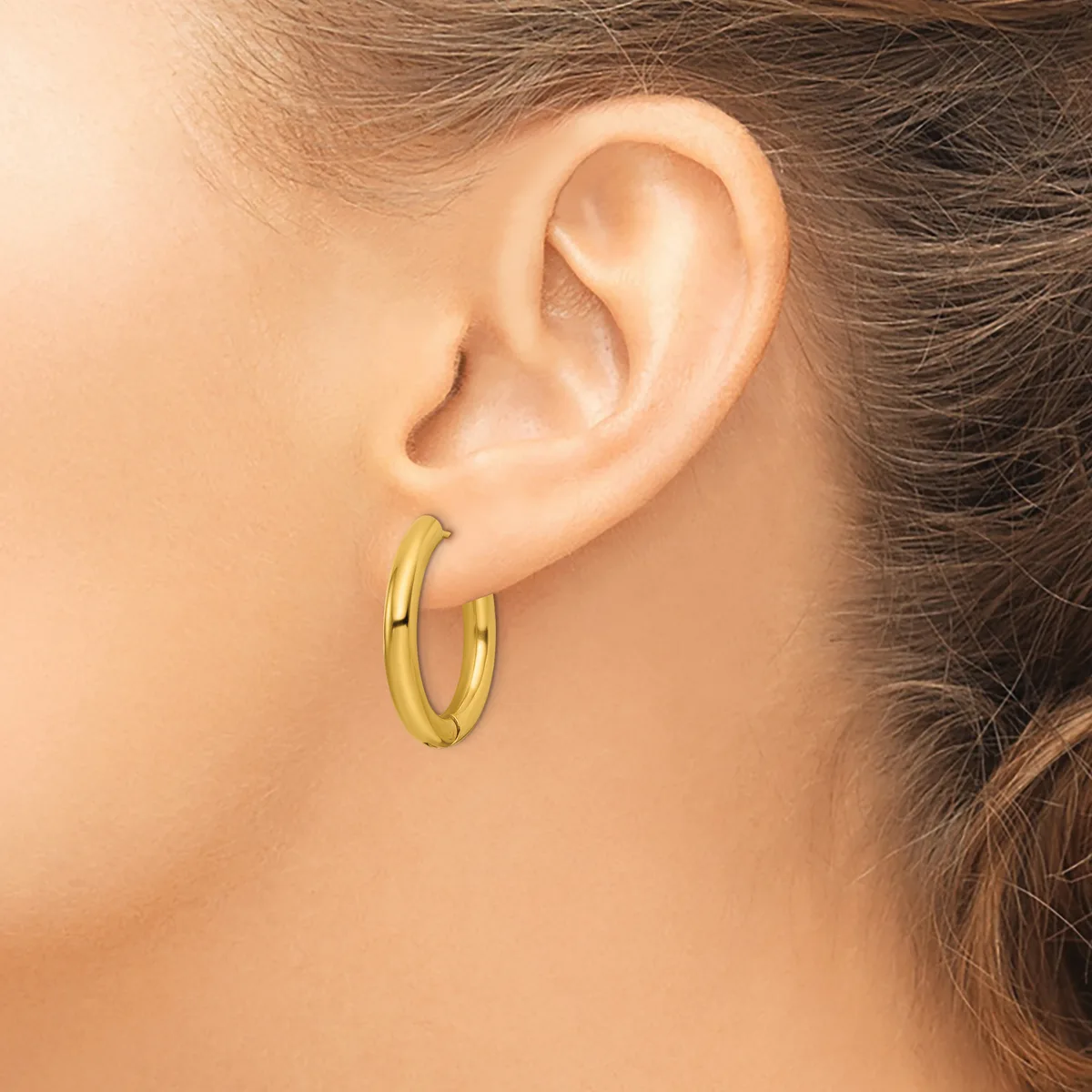 A woman wearing a pair of polished gold-toned hoop earrings, shown from the side profile of her face and ear.