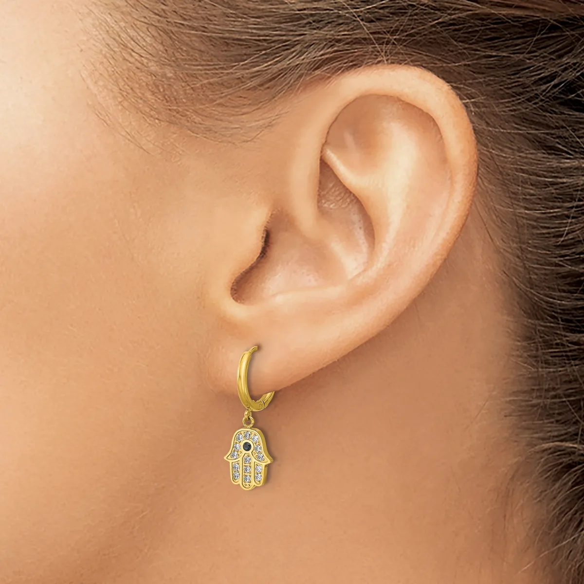 A person wearing a gold hoop earring with a dangling hamsa hand charm adorned with small white stones and a blue stone in the center.
