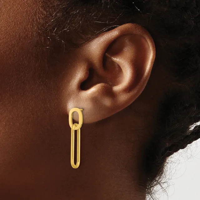 A gold paperclip-shaped earring hangs from the earlobe of a person with brown skin and dark hair.
