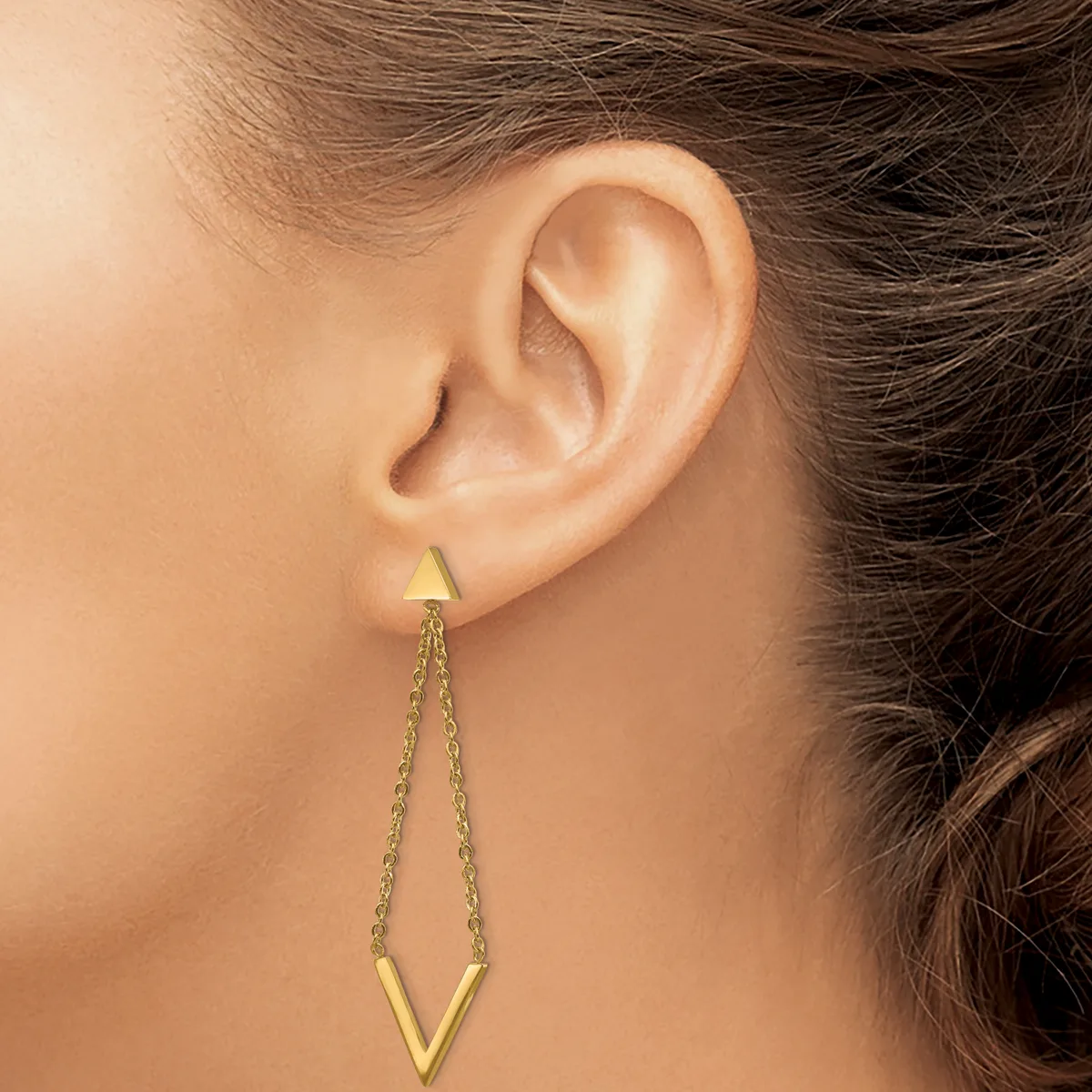 A person wearing gold-toned dangle earrings with a small triangular stud at the top connected by chain links to a larger inverted V-shaped pendant below.